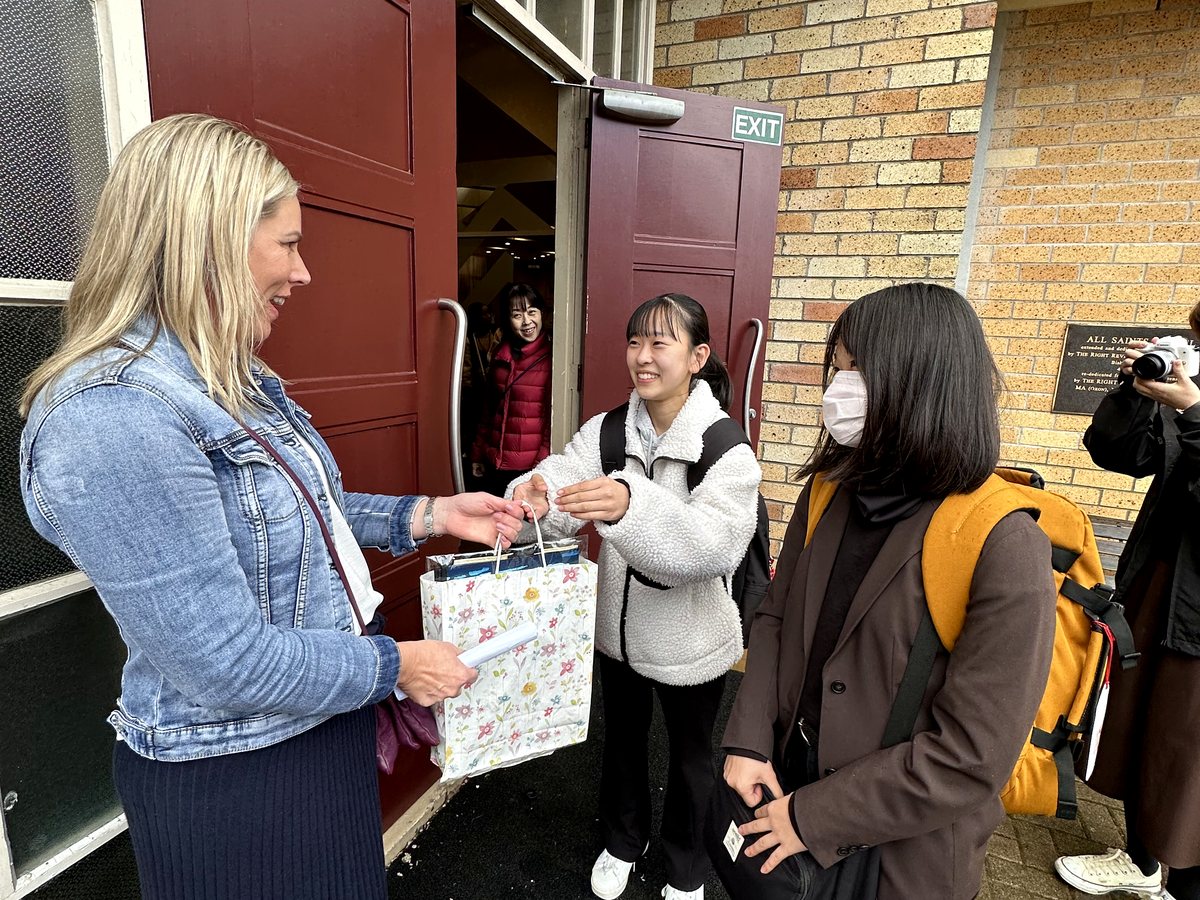 A warm, candid outdoor interaction where a blonde woman (staff or host) presents a floral gift bag to a...