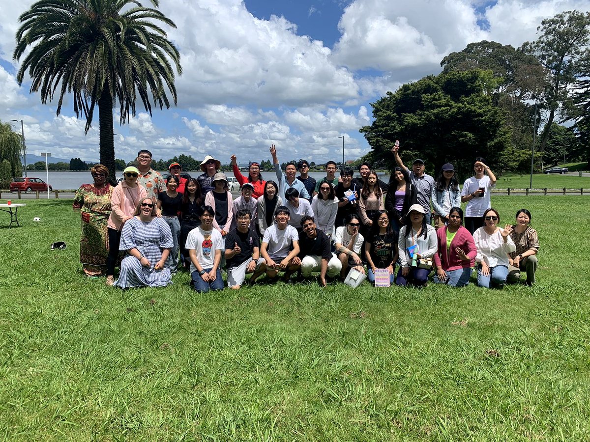 A large group of diverse international students and staff posing together on a grassy park lawn near Lake...