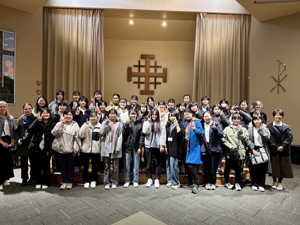 A large group of international students and staff posing together indoors on a stage, with many students...