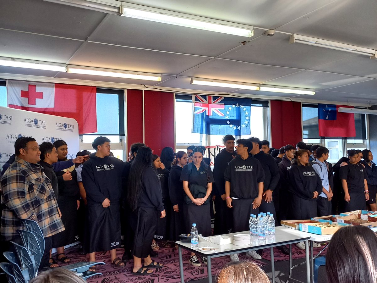 A group of Pasifika students and community members gathered indoors wearing 'AIGA TASI' branded apparel...