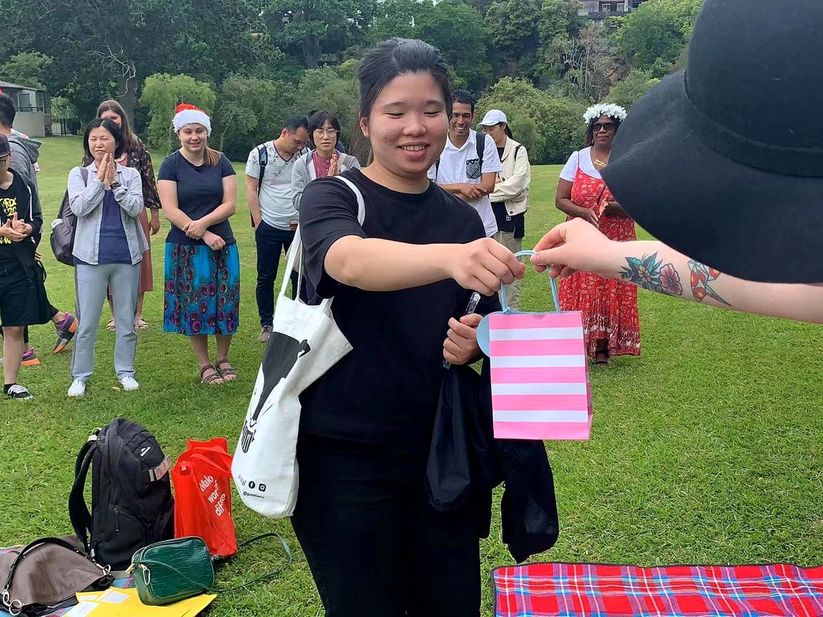 A smiling Asian female student receives a pink striped gift bag from a staff member or student outdoors...