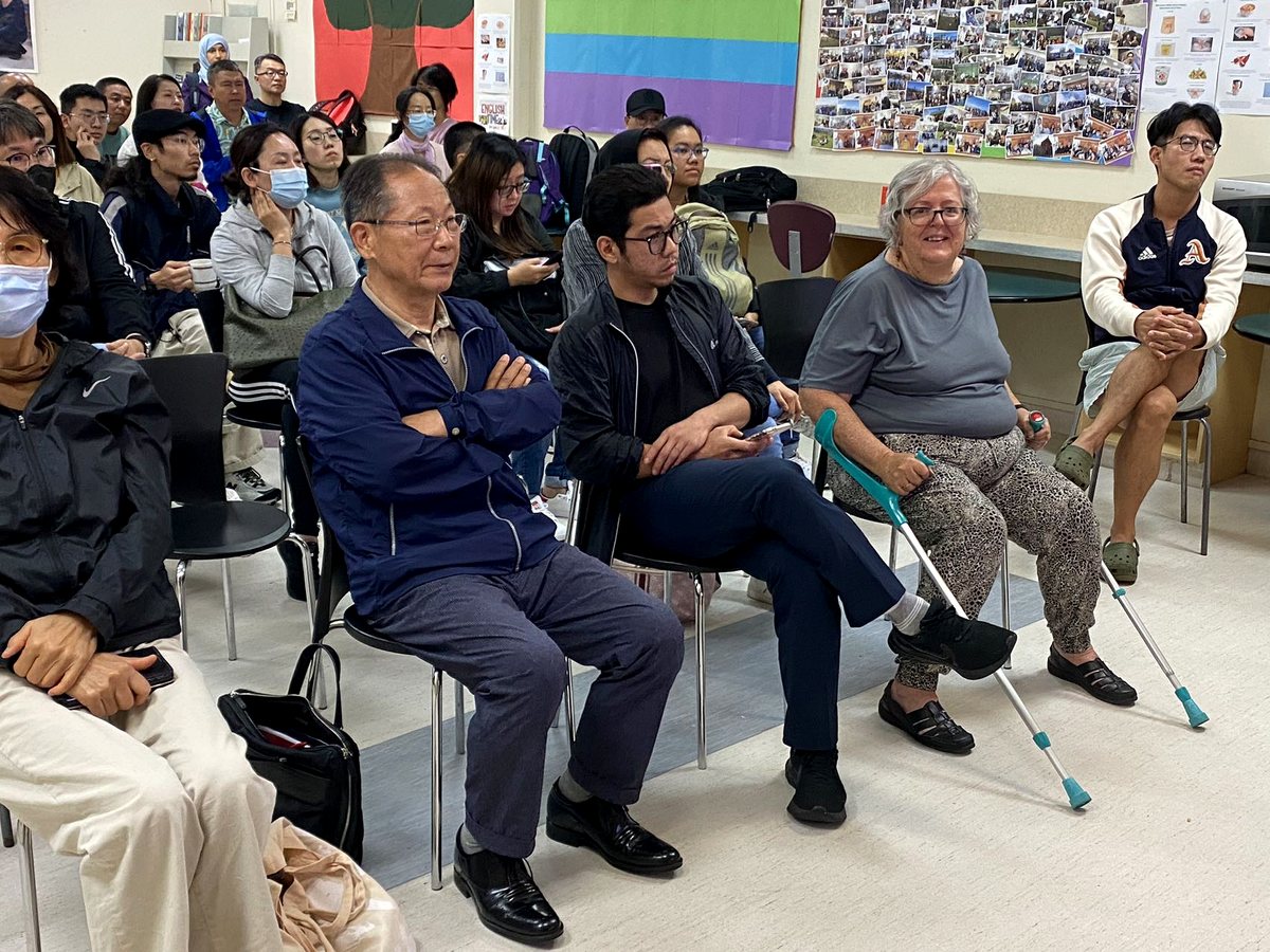 A diverse group of adult students and community members sitting attentively in a classroom or common room...