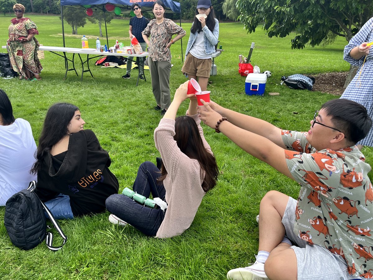 Diverse international students enjoying an outdoor picnic and social activity on a park lawn in Hamilton,...